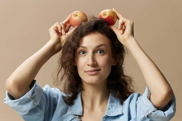 Lovely funny curly beautiful female in jeans casual shirt with
apple overhead as ears posing isolated on over beige pastel
background healthy food natural ecofriendly products concept copy
space