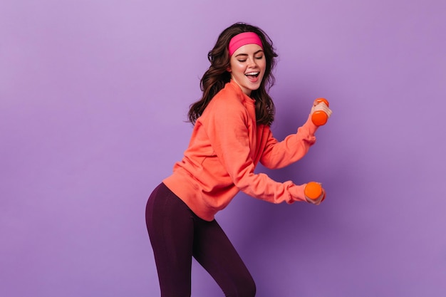 Photo lovely girl in black leggings and orange sweatshirt does aerobics with dumbbells