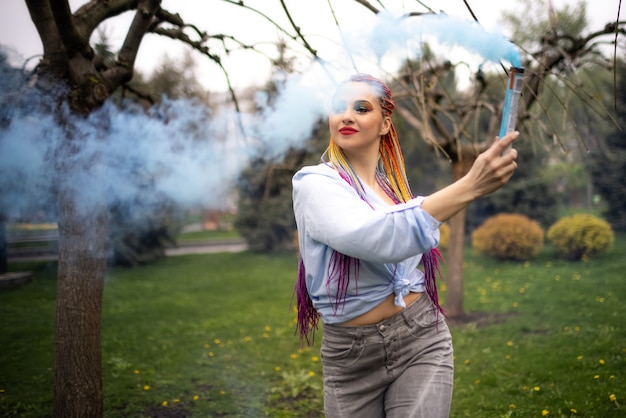 A lovely girl in a bluish shirt with expressive makeup and colored African braids. Posing and spinning in the dense sky-blue artificial smoke in a blooming park.