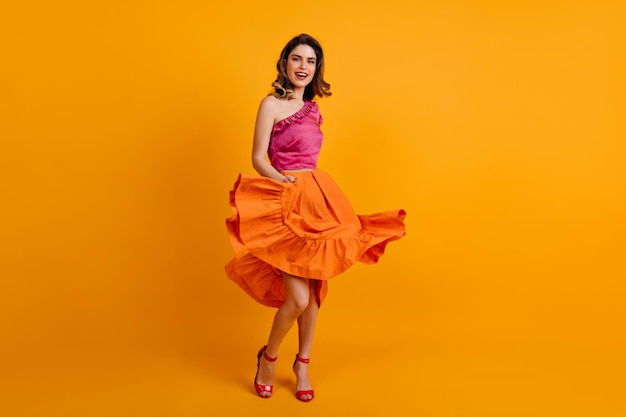 Lovely girl in colorful attire dancing with smile Refined caucasian woman playing with her orange skirt