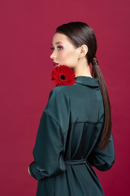 Lovely girl wearing green dress standing isolated over red background posing with a herbera flower