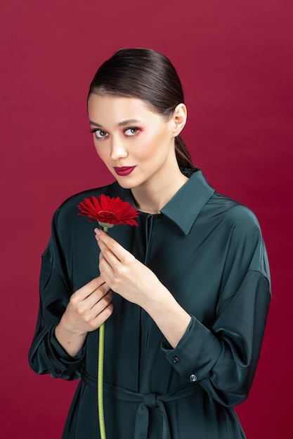 Photo lovely girl wearing green dress standing isolated over red background posing with a herbera flower