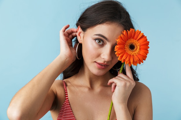 Bella ragazza che indossa abiti estivi in piedi isolato sul blu, in posa con un fiore di herbera