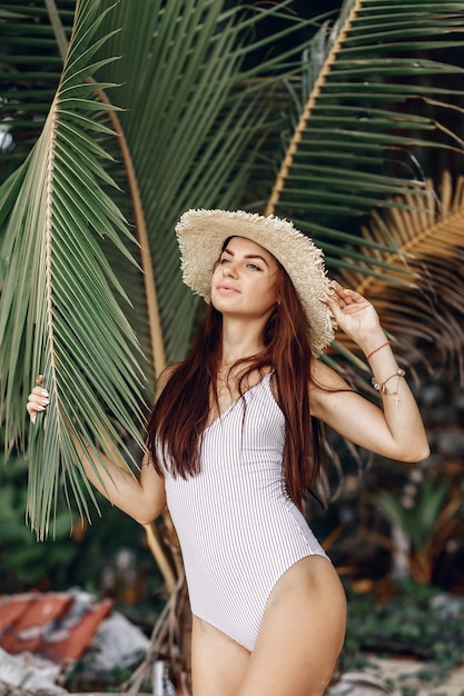 Lovely girl in a white bodysuit and a straw hat is posig next to large palm leaves on a jungle background. Summer concept