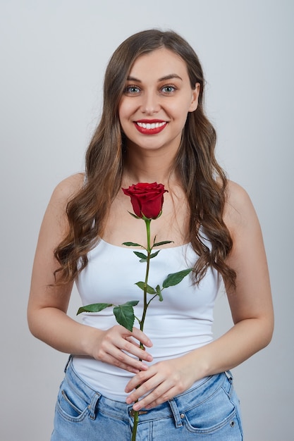 흰색 티셔츠에 사랑스러운 소녀는 회색 벽에 웃고, 빨간 장미를 들고있다. 행복한 발렌타인 데이.