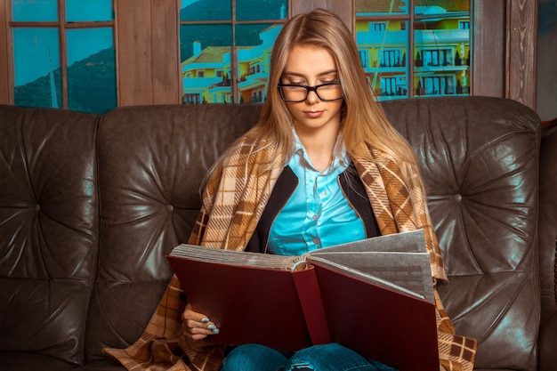 Lovely girl with beautiful long hair sitting at a large window on a brown leather couch, a blanket hiding and seriously looks in the red book