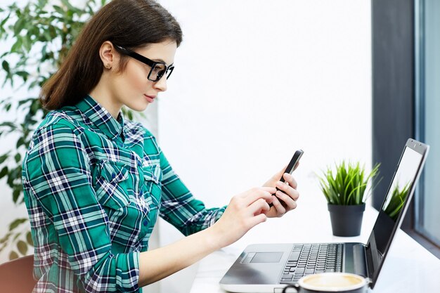 Lovely girl with black hair wearing shirt and eyeglasses sitting in cafe with laptop and cup of coffee, freelance concept, working process, scanning QR-code, online shopping.