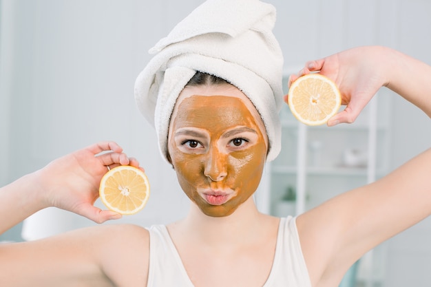 Lovely girl with brown facial mask holding a slice of lemon near her face and smiling. Photo of girl receiving spa treatments. Beauty and Skin care concept