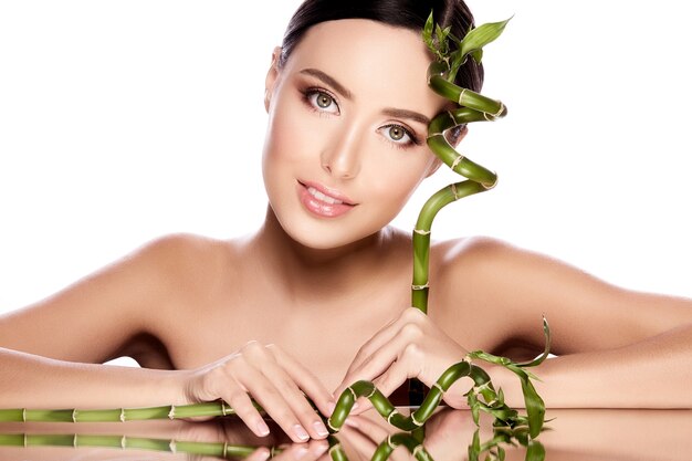 Lovely girl with brown hair fixed behind, clean fresh skin, big eyes and naked shoulders posing with green plant at white  wall, skin treatment concept.