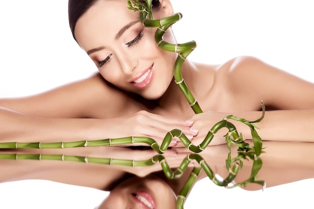 Lovely girl with brown hair fixed behind, clean fresh skin, big eyes and naked shoulders posing with green plant at white  wall, skin treatment concept.