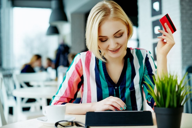 Lovely girl with light hair wearing colorful shirt sitting in cafe with tablet, credit card and cup of coffee, freelance concept, online shopping.