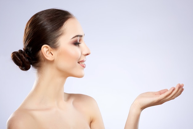 Lovely girl with nude make up posing at grey studio background, beauty photo concept, perfect skin, holding product and looking at it.