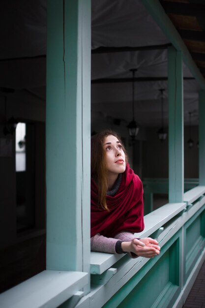 Lovely girl with a red scarf in an autumn coat stands on the terrace of a cafe in the city.