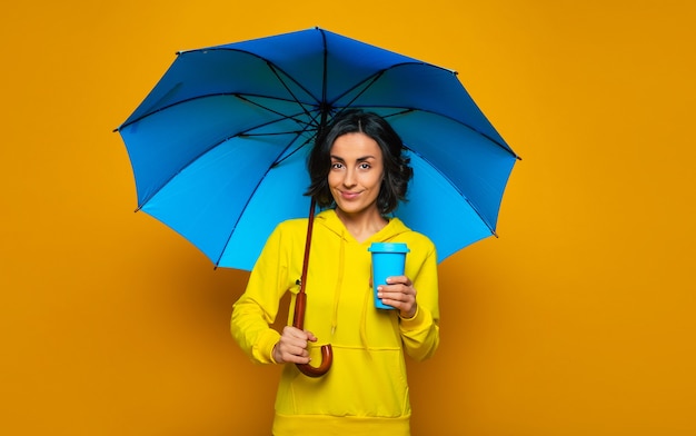 lovely girl with smile on her face under a blue umbrella, dressed in a yellow hoodie, holding blue thermal mug in her left hand, ready for the rainy day.