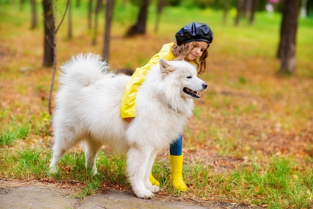 Lovely girl in yellow rubber boots and rain coat on a walks, plays with a beautiful white samoyed dog in the autumn park.