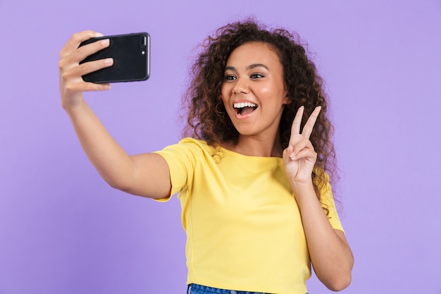 Bella ragazza africana felice che indossa abiti casual in piedi isolata sul muro viola, facendo un selfie, mostrando un gesto di pace