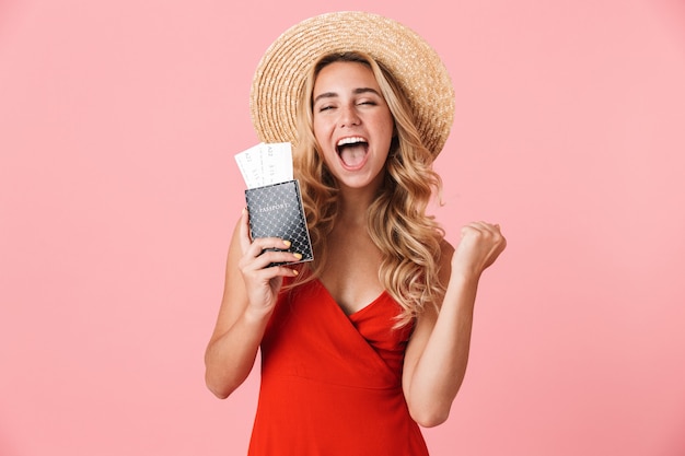 Lovely happy young blonde woman wearing summer dress standing isolated over pink wall, showing passport with flight tickets, celebrating