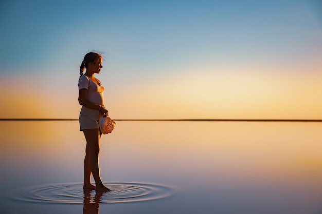 Lovely happy young girl walking along the mirror pink salt lake enjoying the warm evening sun looking at the fiery sunset and her reflection