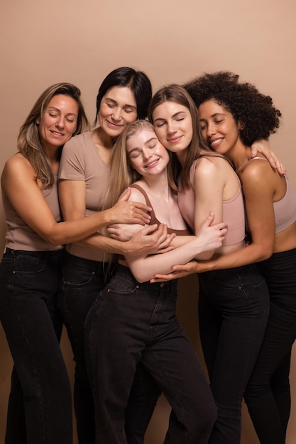 Lovely interracial dark and blonde ladies in tops and pants with closed eyes huddle together on beige background