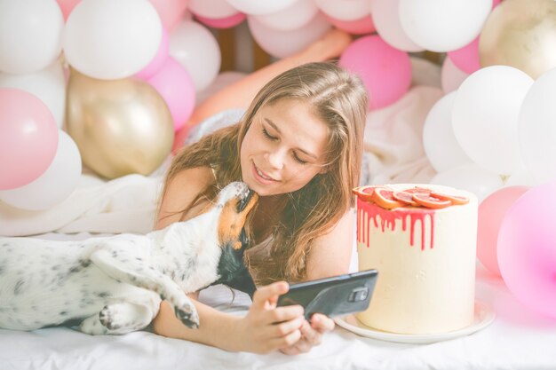 Photo lovely lady in pajama making selfie in her bedroom using phone and kiss  her dog.