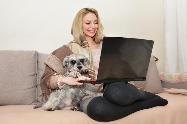 Lovely lady smiles and hugging a cute dog on the couch while watching movies on a laptop