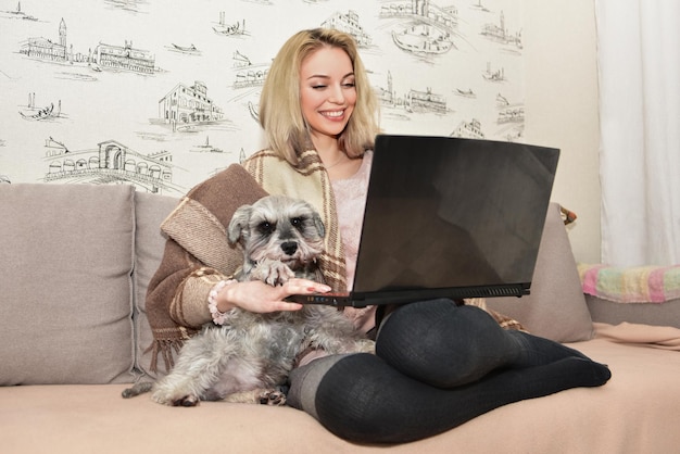 Lovely lady smiles and hugging a cute dog on the couch while watching movies on a laptop