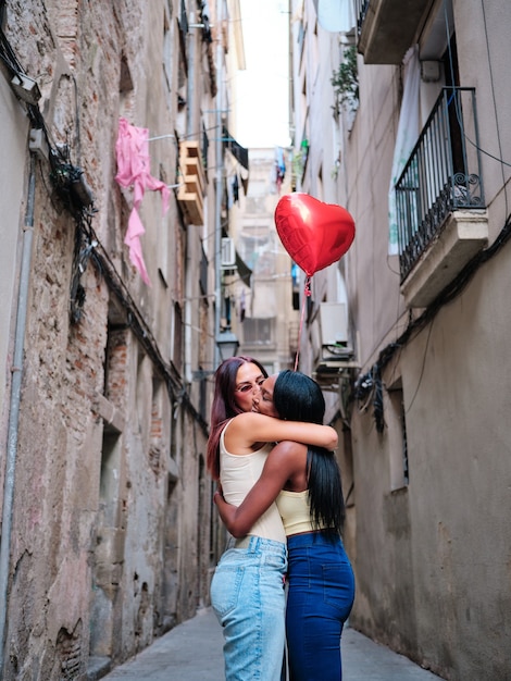 Lovely lesbian couple hugging while holding a red heart-shaped balloon outdoors on the street. LGBT and Valentine's day concept.