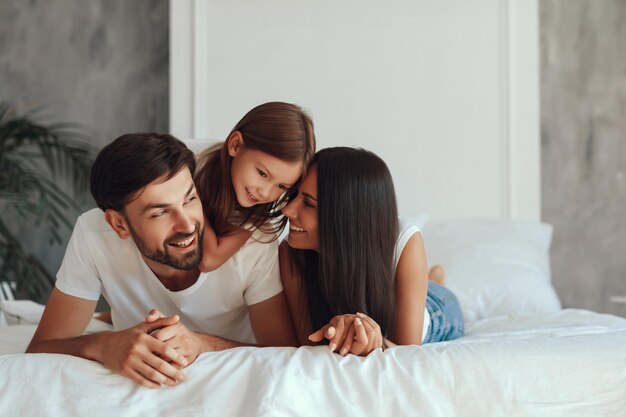 Lovely little girl snuggling between her mom and dad while laying on a bed with white linen