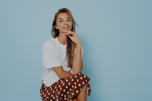 Lovely long haired woman in stylish outfit ready for evening walk with boyfriend or friends, looking cute and flirty while sitting on chair against blue background. Female beauty concept