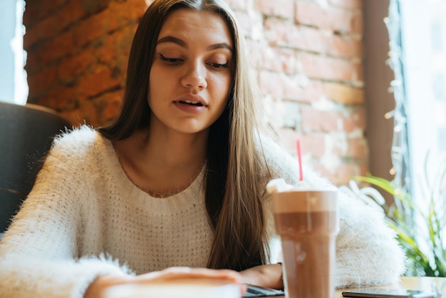 Lovely longhaired girl freelancer in a white sweater sits in a cafe works and drinks a fragrant cappuccino