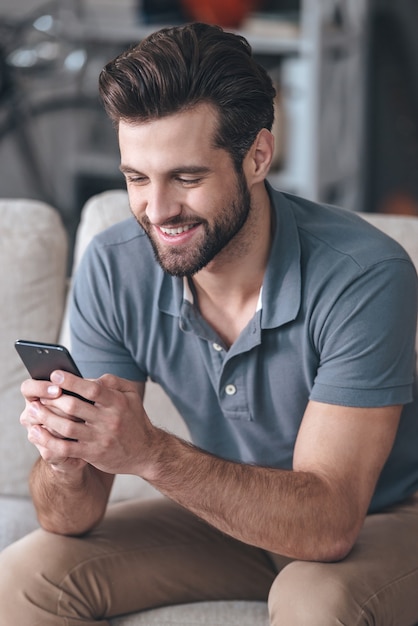 Bel messaggio per un amico. bel giovane che guarda il suo smartphone con un sorriso mentre è seduto sul divano di casa