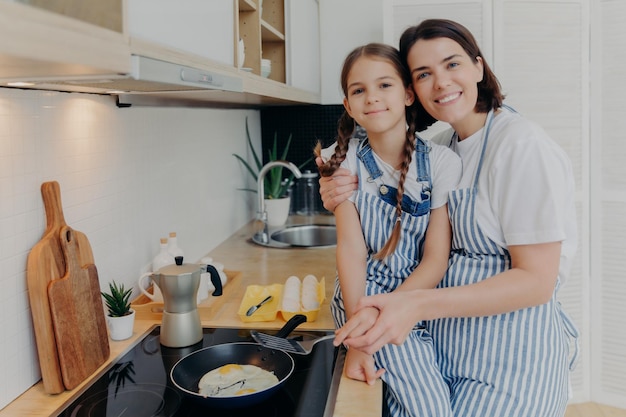 Madre e figlia adorabili in grembiuli abbracciano e sorridono allegramente friggono le uova sul fornello moderno in cucina usano la padella preparare una gustosa colazione famiglia bambini maternità e concetto di cucina