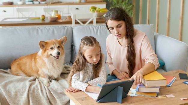 A Lovely Mother Helps to Her Cheerful Daughter Do Homework. Girl Takes a Remotely Education. Doing Homework With a Tablet.