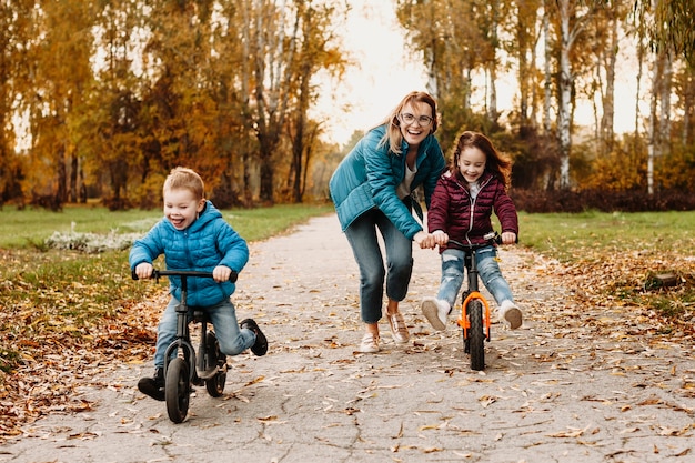 Прекрасная мать играет со своими детьми, склоняя свою дочь кататься на велосипеде, в то время как ее маленький сын идет вперед со смехом на велосипеде.