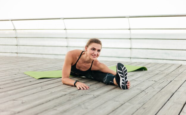 Lovely muscular sportswoman in sportswear doing leg stretching while sitting in twine on beach terrace. Outdoor sports