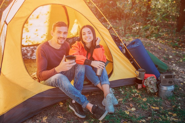 Lovely picture of couple sitting together in tent and look. They smile. People hold thermo cups. There is backpack with equipment.