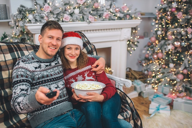 Lovely picture of young couple sitting together and looking on camera. He holds remote control. She has bowl of popcorn in hands. They smile. People are in decorated room.