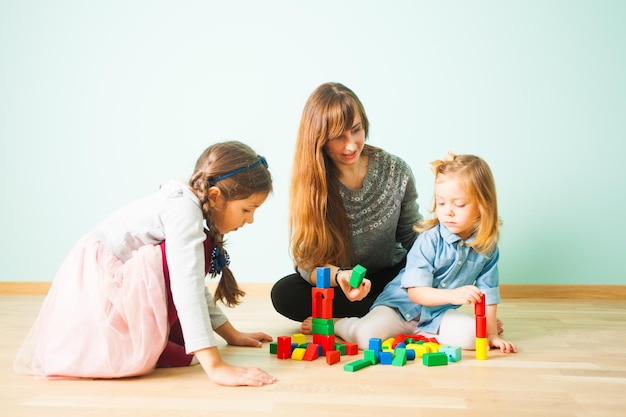 Lovely portrait of young attractive mother with long dark hair and her adorable daughters playing with wooden blocks play set. Woman proposing green cube to girl building colorful tower