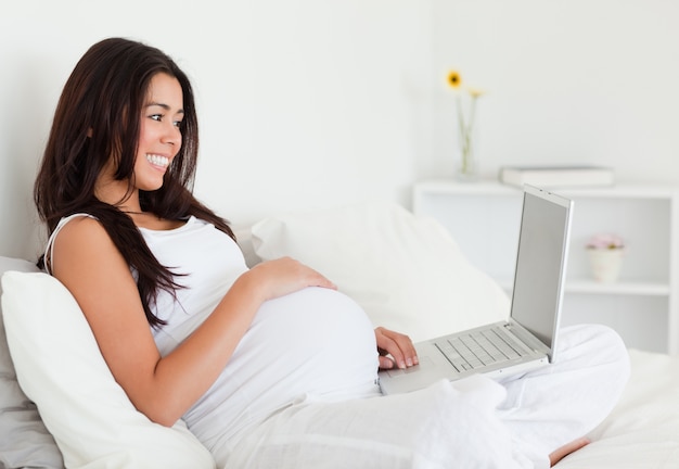 사랑스러운 임신 한 여자는 침대에 누워있는 동안 그녀의 노트북으로 편안한