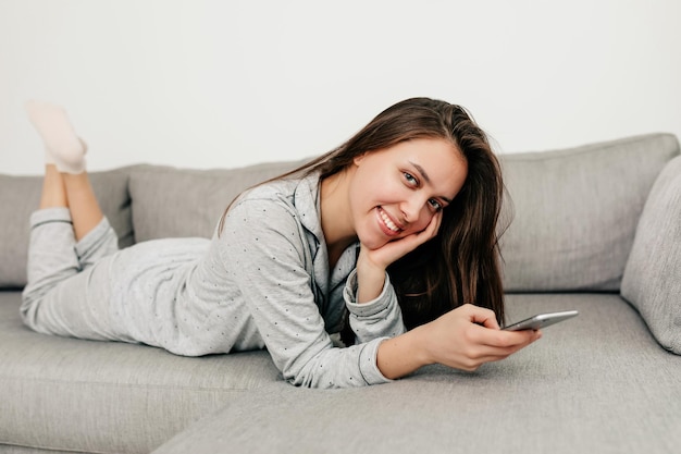 lovely pretty girl with long dark hair is wearing home suit lying on sofa and smiling at camera with smartphone Indoor portrait of happy girl is relaxing at home