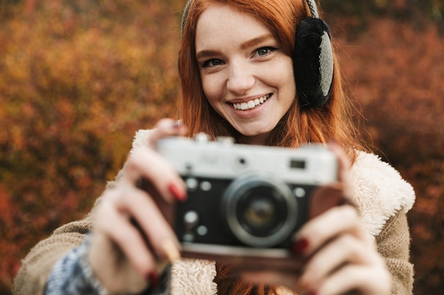 Lovely redheaded young girl listening to music, holding photo camera while standing outdoors