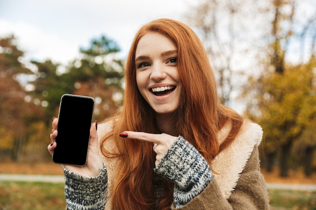 Photo lovely redheaded young girl listening to music while sitting on a bench, using mobile phone, showing blank screen mobile phone