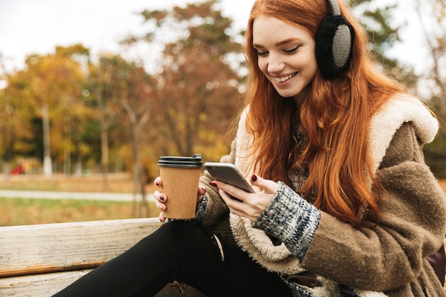휴대 전화를 사용하여 벤치에 앉아있는 동안 헤드폰으로 음악을 듣고 사랑스러운 빨간 머리 어린 소녀