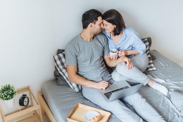 Lovely romantic couple going to kiss look at each other as sit on bed use laptop computer for watching films drink coffee enjoy togetherness and relaxation in bedroom Relationship concept