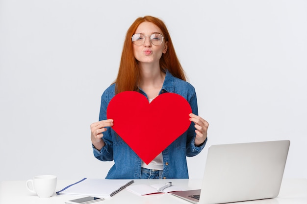 Lovely, romantic cute redhead girl folding lips in mwah, send air kiss and holding big red valentines card, showing her love and passion, congratulate girlfriend with anniversary, stand near laptop