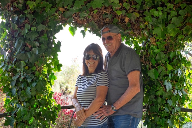 Прекрасная пожилая пара в джинсах и солнцезащитных очках обнимается, расслабляясь в саду, глядя в камеру