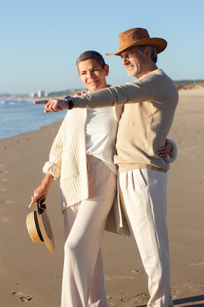 Lovely senior couple standing at the beach, looking at seascape. Bearded man in cowboy hat pointing at something while short-haired lady with straw hat in hand hugging him. Leisure, love concept