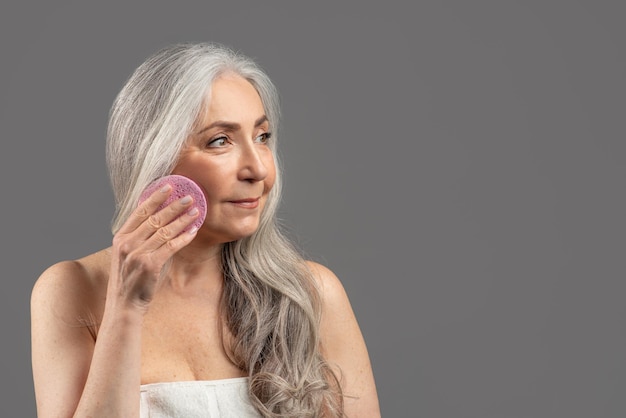 Lovely senior woman with beautiful long hair holding cosmetic sponge near her face on grey studio