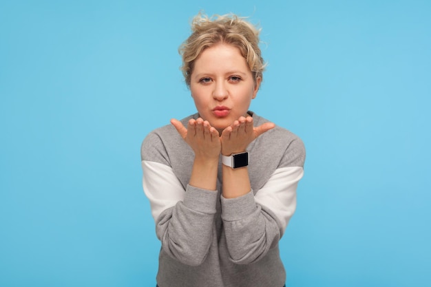 Lovely sensual woman with short curly hair in casual sweatshirt sending air kiss to camera, expressing affection and fondness, romantic feelings, love. indoor studio shot isolated on blue background