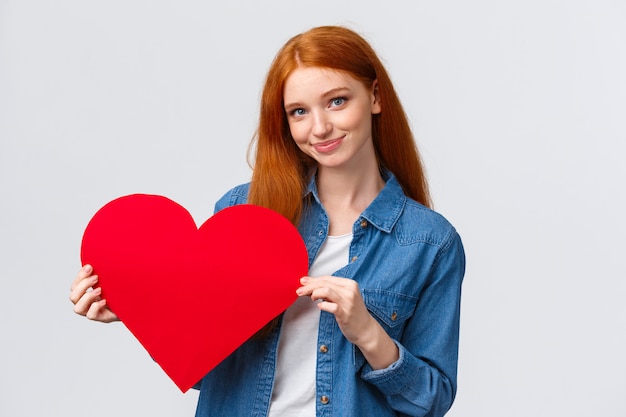 Adorabile e timida ragazza carina rossa confessa la simpatia, regali nel giorno di san valentino, cuore rosso fatto a mano, macchina fotografica sciocca e tenera sorridente, esprimi amore e sentimenti, in piedi bianca